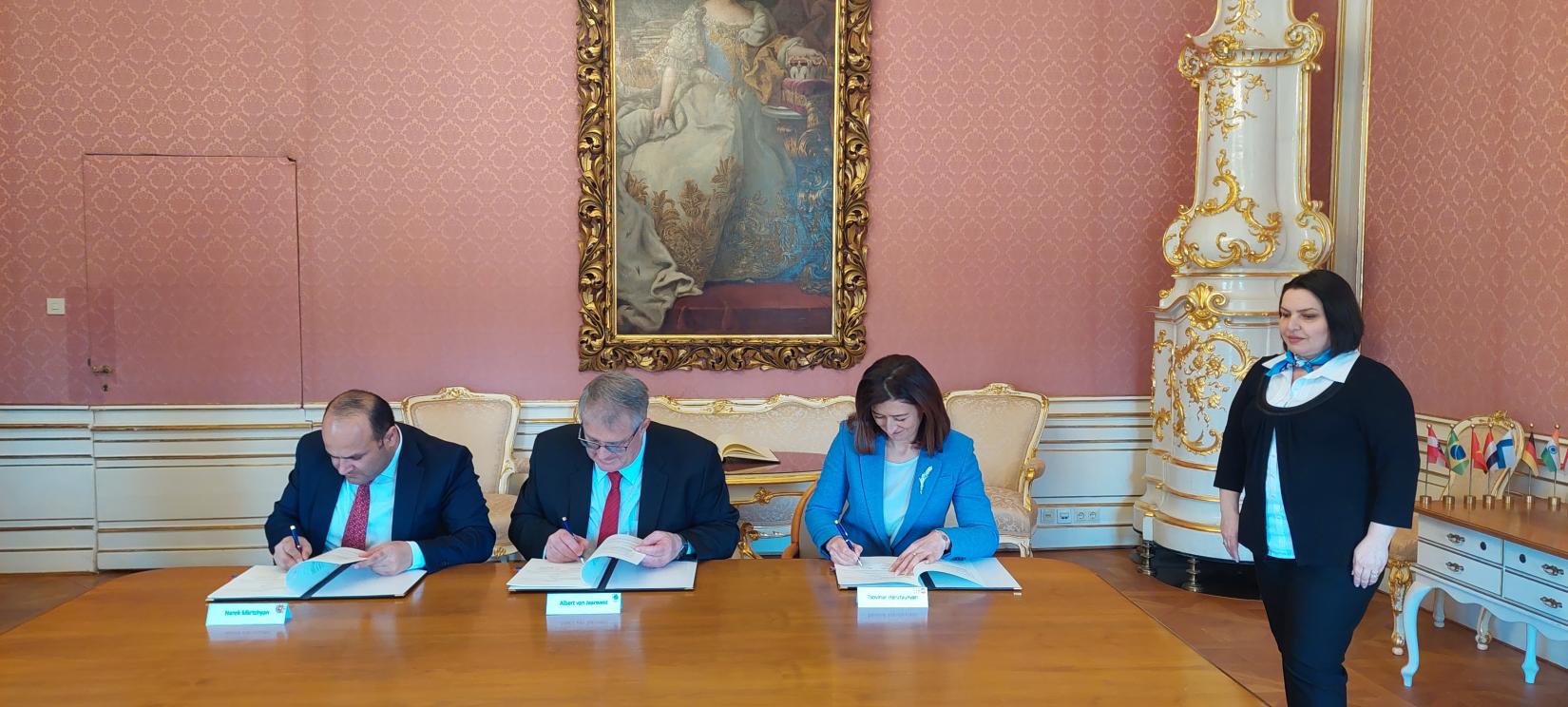 Three people (two men and a woman) are signing the Memorandum of Understanding and a woman is standing next to them.