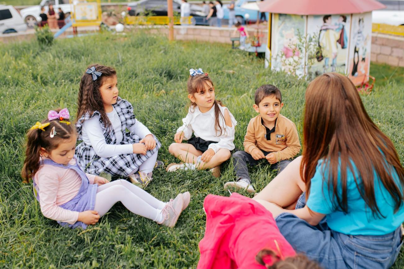 The UNICEF staff member talks to the children (including three girls and a boy) sitting on the grass.